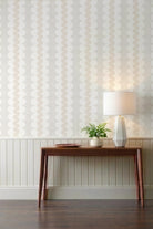 A styled interior scene showing a wall with vertical wavy patterned wallpaper, a wooden console table, a lit table lamp, and a small potted plant against a white wainscoted wall.