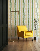 A styled interior scene featuring a bright yellow armchair in front of a wall with repeating vertical wavy stripes in green and cream. A tall brass floor lamp stands to the left, and a small side table with a vase of branches sits to the right.