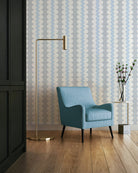 A styled interior scene featuring a light blue upholstered armchair in front of patterned wallpaper, with a gold floor lamp and a small side table holding a vase with branches.