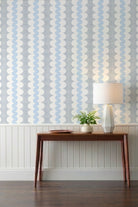 A styled interior scene showing patterned wallpaper with vertical wavy stripes in light blue and white, installed above white wainscoting. A wooden console table, a white table lamp, and a small potted plant are placed against the wall on a dark wood floor.