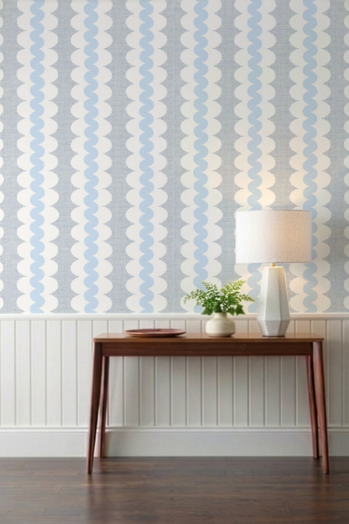 A styled interior scene showing patterned wallpaper with vertical wavy stripes in light blue and white, installed above white wainscoting. A wooden console table, a white table lamp, and a small potted plant are placed against the wall on a dark wood floor.