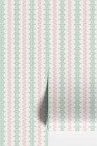 Striped wallpaper with a curvy stripe in soft pastel pink, on a stripe of white oval shapes, with a green square grid in the background. Shown as two wallpaper rolls on a wall.