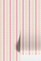 A close-up of wallpaper or fabric with a repeating vertical pattern of pink wavy lines over a nectarine and white scalloped background, with a folded corner showing a light gray underside.