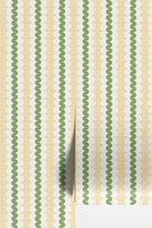 Striped wallpaper with a curvy stripe in olive green, on a stripe of white oval shapes, with a yellow square grid in the background. Shown as two wallpaper rolls on a wall.