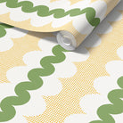 A roll of wallpaper partially unrolled, showing a retro pattern with olive green wavy lines, mustard yellow square grid with  white scalloped oval shapes.