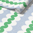 A roll of wallpaper with a retro pattern of green wavy lines, blue square dots, and white oval shapes, shown partially unrolled on a white surface.