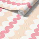 A roll of wallpaper partially unrolled, showing a retro pattern with pink wavy lines, peach square dots and white ovals on a white background.