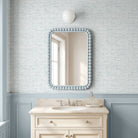 A bathroom vanity scene with a rectangular wall mirror featuring a light blue beaded frame, mounted on a light blue and white patterned wallpaper above a cream-colored cabinet with a marble sink.