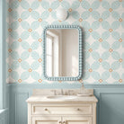 A styled bathroom interior featuring a cream vanity with an undermount sink, a beaded light blue mirror frame, and wallpaper with a repeating teal and beige floral pattern above a light blue wainscoted wall.