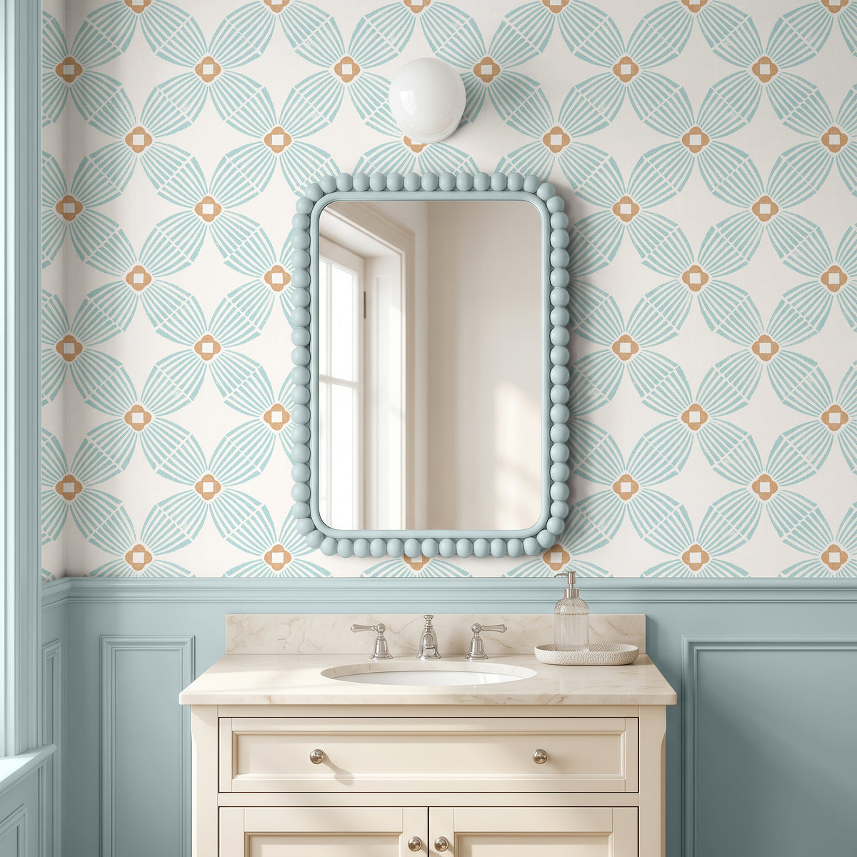 A styled bathroom interior featuring a cream vanity with an undermount sink, a beaded light blue mirror frame, and wallpaper with a repeating teal and beige floral pattern above a light blue wainscoted wall.