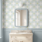 A styled bathroom interior featuring a cream vanity with a marble sink, a light blue beaded mirror frame, and wallpaper with a repeating floral pattern in light blue and olive green.
