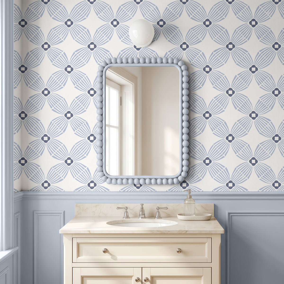 A bathroom vanity scene with a cream-colored pedestal sink and marble countertop beneath a beaded mirror, set against wallpaper with a repeating blue and white geometric pattern and blue wainscoting.