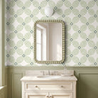 A bathroom vanity scene with a rectangular mirror featuring a beaded frame, mounted on wallpaper with a green and white geometric pattern. Below the mirror is a white pedestal sink with a marble countertop and chrome faucet.
