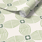 A roll of wallpaper with a repeating geometric pattern in sage green and dark green on a warm off-white background, shown partially unrolled.