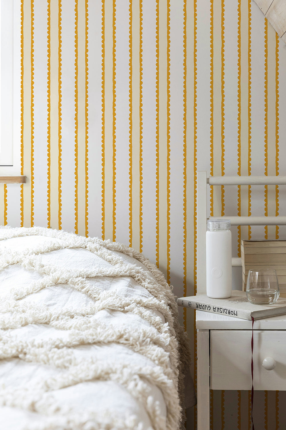 Yellow hand drawn scallops and white vertically striped wallpaper shown in a styled bedroom setting with a bed, bedside table, and white bedding.