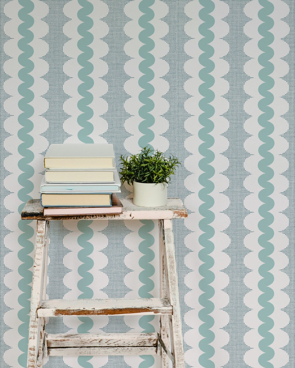 A styled interior scene showing a distressed white wooden stool with a stack of books and a small potted green plant in front of a wall covered in vertical wavy stripes in pale aqua and white.