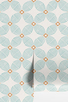 A close-up of wallpaper rolls with a repeating geometric floral pattern in light aqua blue and warm tan on an off-white background.