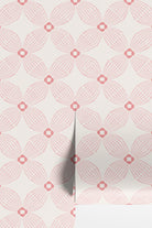 Pink and white geometric wallpaper rolls with a repeating fan-like pattern and small diamond medallions, shown with a folded corner revealing a white surface underneath.