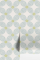 A close-up of wallpaper with a repeating geometric floral pattern in pale blue-gray and muted yellow-green on an off-white background.