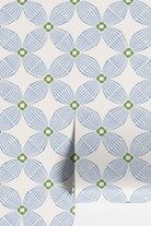 A close-up of wallpaper rolls with a repeating geometric pattern of blue and green stylized flowers on an off-white background.