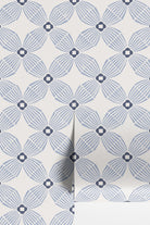 A close-up of wallpaper with a repeating geometric floral pattern in muted blue-gray on an off-white background, with a folded corner showing the texture and layering.