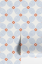 A close-up of wallpaper rolls with a repeating geometric floral pattern in light  blue and blaze orange on an off-white background.