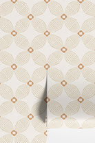 A close-up of wallpaper with a repeating geometric floral pattern in warm beige and tan on an off-white background, with a folded corner showing the surface depth.