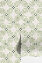 A close-up of wallpaper with a repeating geometric floral pattern in sage green and gray on an off-white background, with a folded corner showing the reverse side.