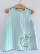 A sleeveless light green and white vertically striped baby dress with a green scalloped hem and an applique apple pocket design on the front, hanging on a wooden hanger against a white wall.