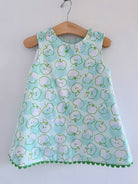 A sleeveless baby dress with an all-over light green and white apple print, hanging on a wooden hanger against a plain white wall.