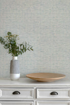 A styled interior scene showing a textured light blue and white wallpaper behind a white wooden dresser. On top of the dresser sits a ribbed white and gray ceramic vase with green eucalyptus stems and a round light wood tray.