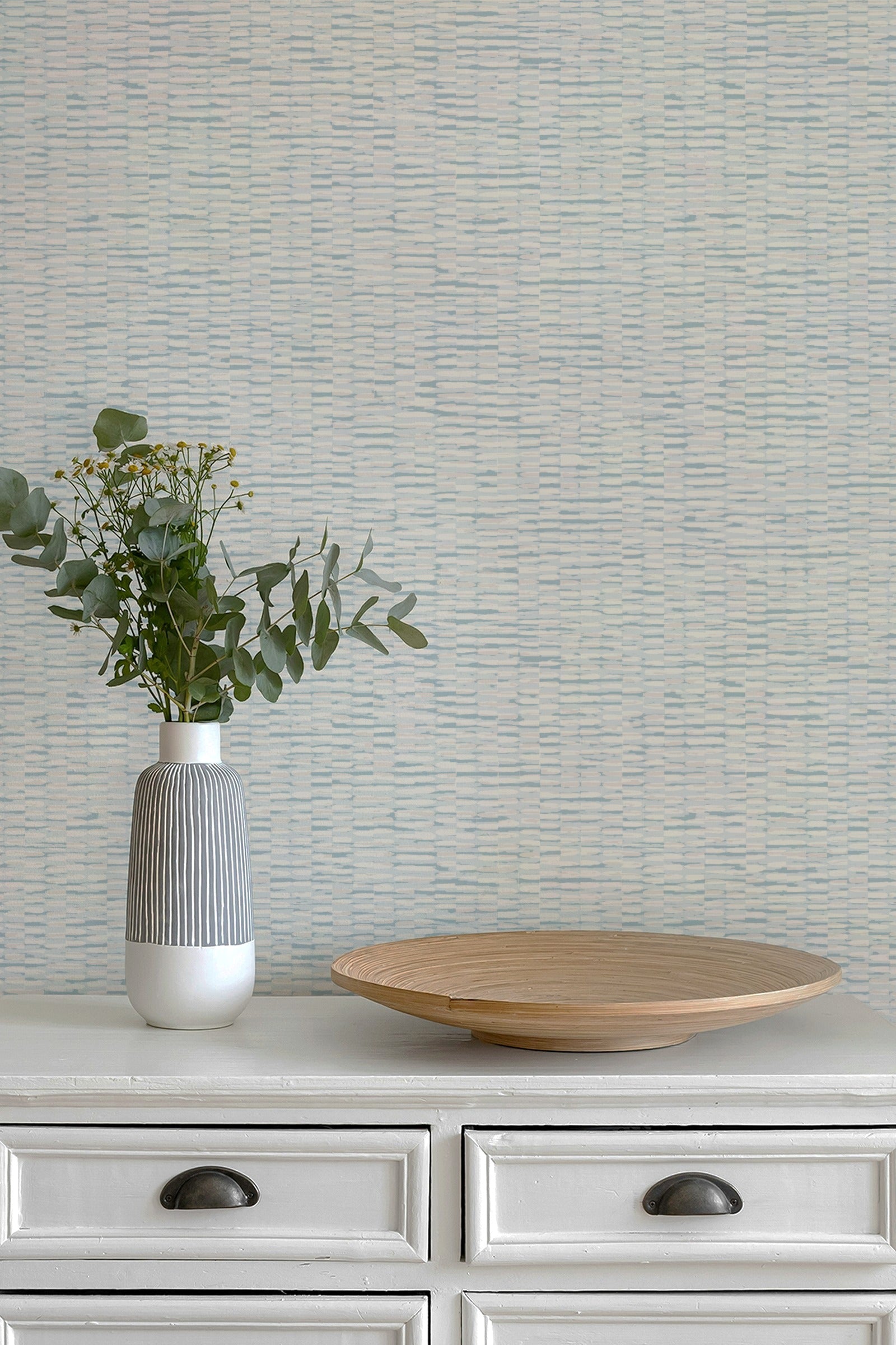 A styled interior scene showing a textured light blue and white wallpaper behind a white wooden dresser. On top of the dresser sits a ribbed white and gray ceramic vase with green eucalyptus stems and a round light wood tray.