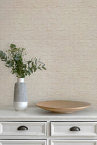 Beige textured wallpaper with a subtle horizontal wavy pattern, styled on a white dresser with a gray and white ceramic vase containing green eucalyptus stems and a round wooden serving platter.