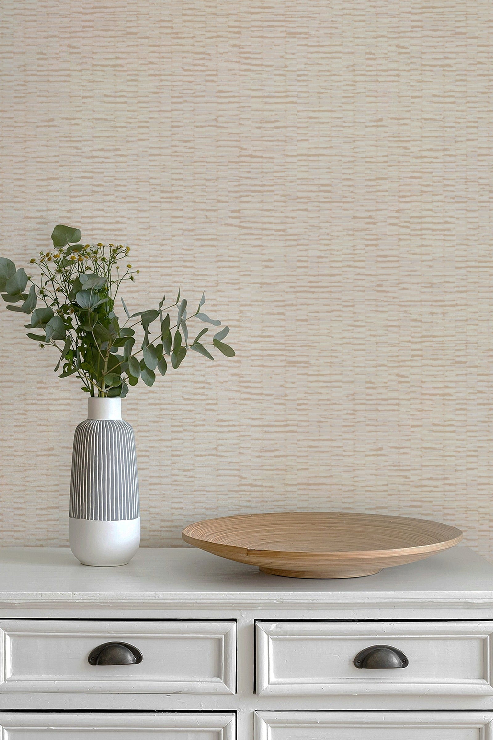 Beige textured wallpaper with a subtle horizontal wavy pattern, styled on a white dresser with a gray and white ceramic vase containing green eucalyptus stems and a round wooden serving platter.
