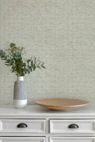 A styled interior scene showing a textured light sage green wallpaper on a wall behind a white dresser. On the dresser sits a ribbed white and gray ceramic vase with green eucalyptus stems and a round light wood tray.