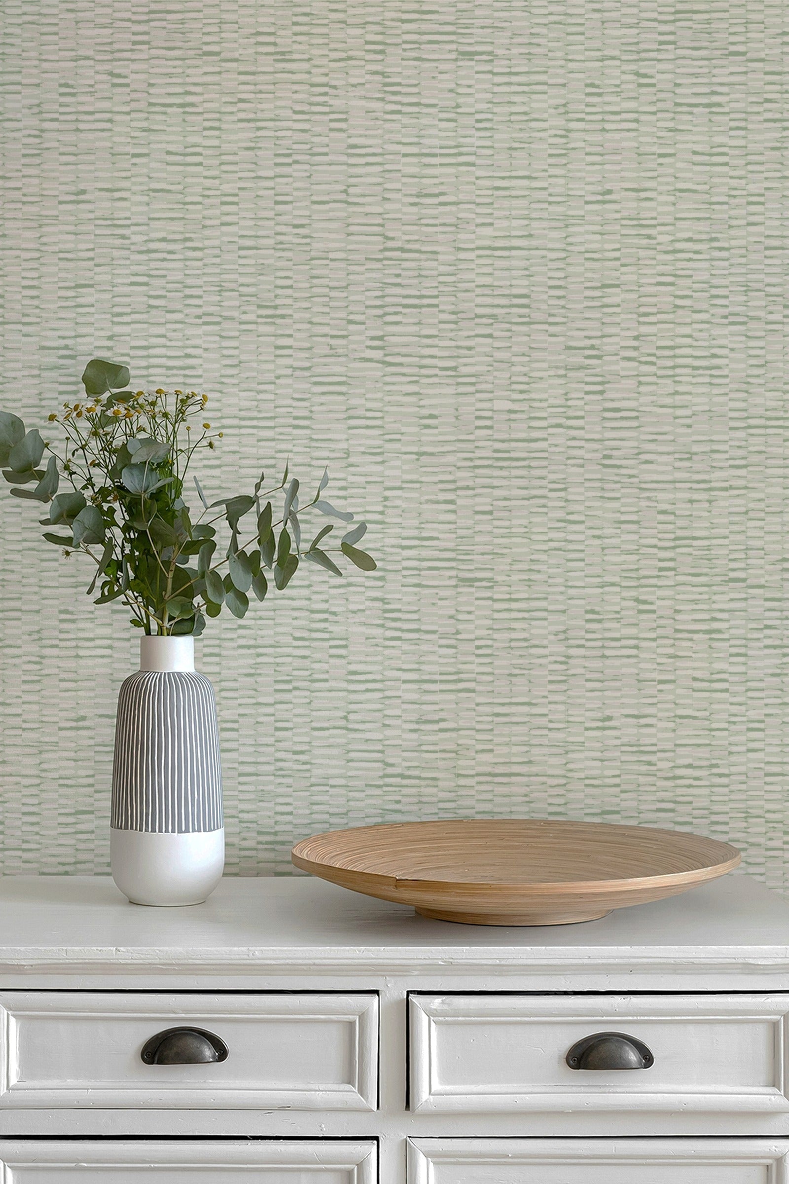 A styled interior scene showing a textured light sage green wallpaper on a wall behind a white dresser. On the dresser sits a ribbed white and gray ceramic vase with green eucalyptus stems and a round light wood tray.
