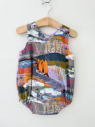 A sleeveless baby romper with a colorful, whimsical landscape print hanging on a wooden hanger against a plain white wall.