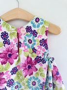 A close-up of a children's sleeveless top or dress with a colorful floral print, hanging on a wooden hanger against a plain white wall.