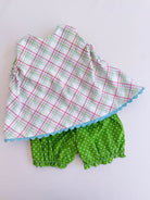 A two-piece baby outfit set featuring a white sleeveless top with a red and green plaid pattern and a light blue scalloped hem, paired with green bloomers with white polka dots, laid flat on a white background.