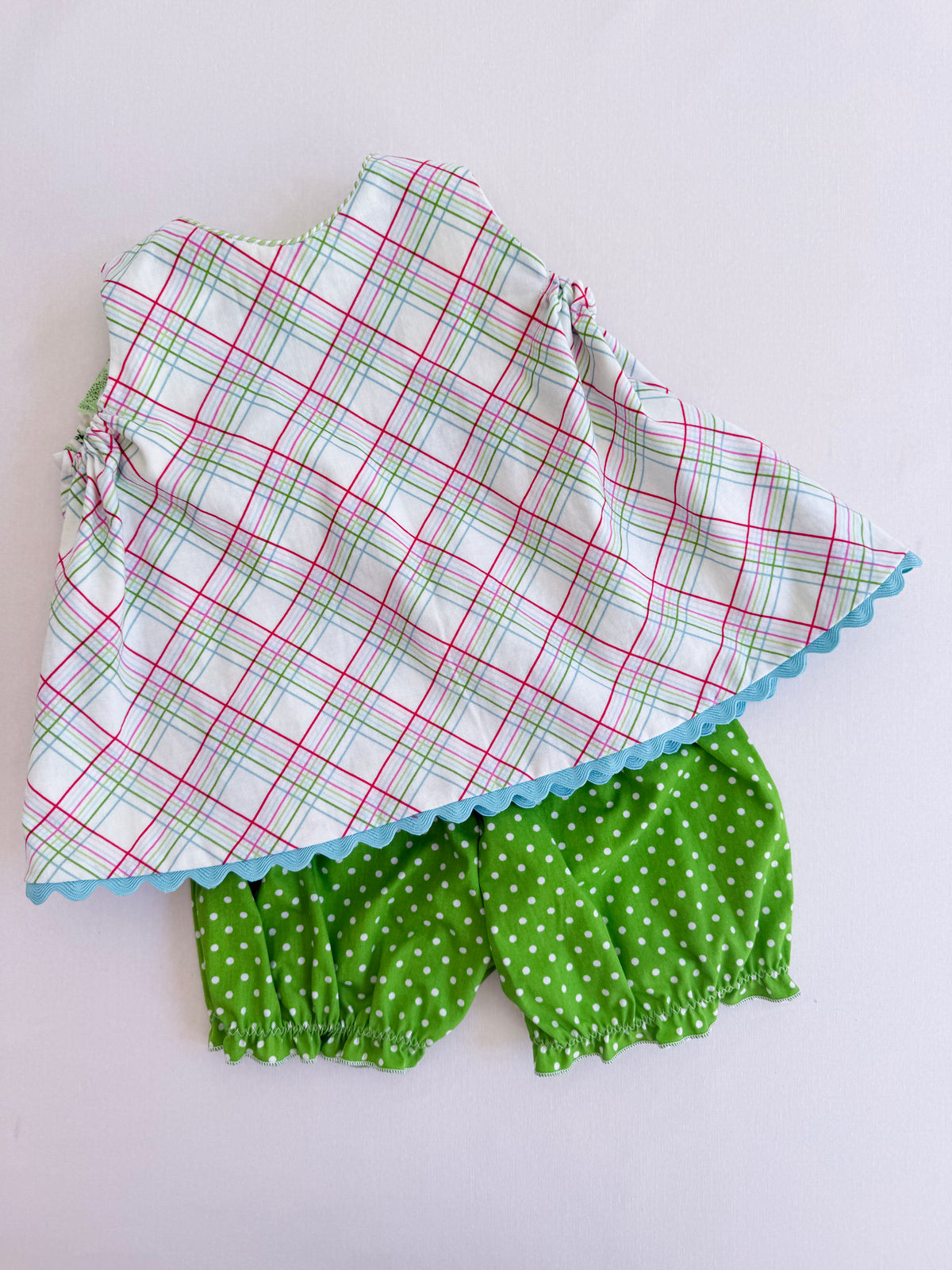 A two-piece baby outfit set featuring a white sleeveless top with a red and green plaid pattern and a light blue scalloped hem, paired with green bloomers with white polka dots, laid flat on a white background.