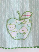 A close-up of a light blue and white patterned fabric with a green applique of an apple featuring smaller apple outlines and a plaid leaf. it is a pocket.