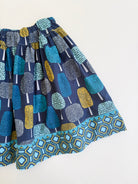 A close up of a gathered child's skirt featuring an all-over abstract tree motifs in teal, blue, yellow, and gray on a navy base, finished with a matching geometric patterned hem.