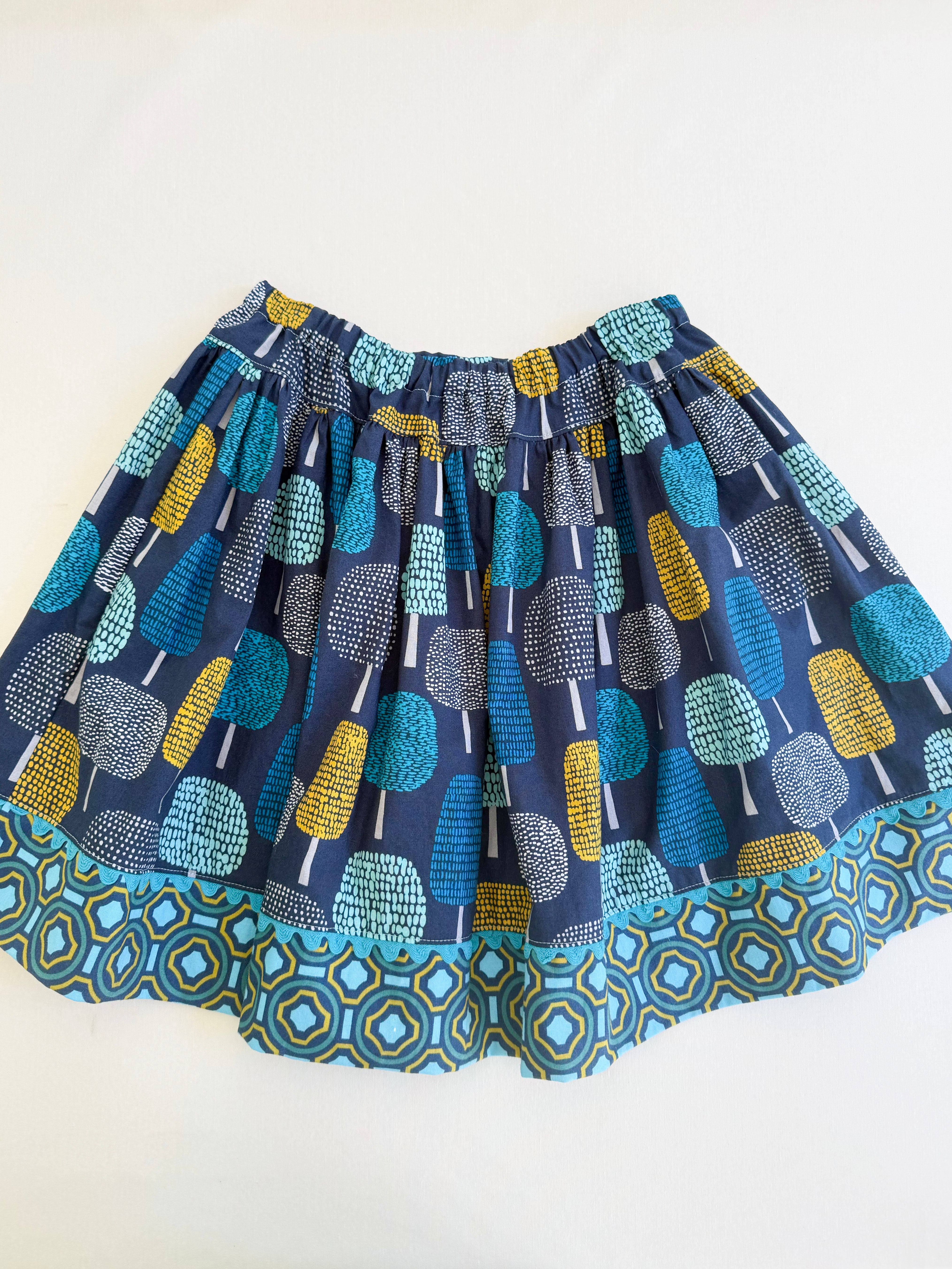 A short, pleated skirt featuring an all-over abstract tree motifs in teal, blue, yellow, and gray on a navy base, finished with a matching geometric patterned hem.